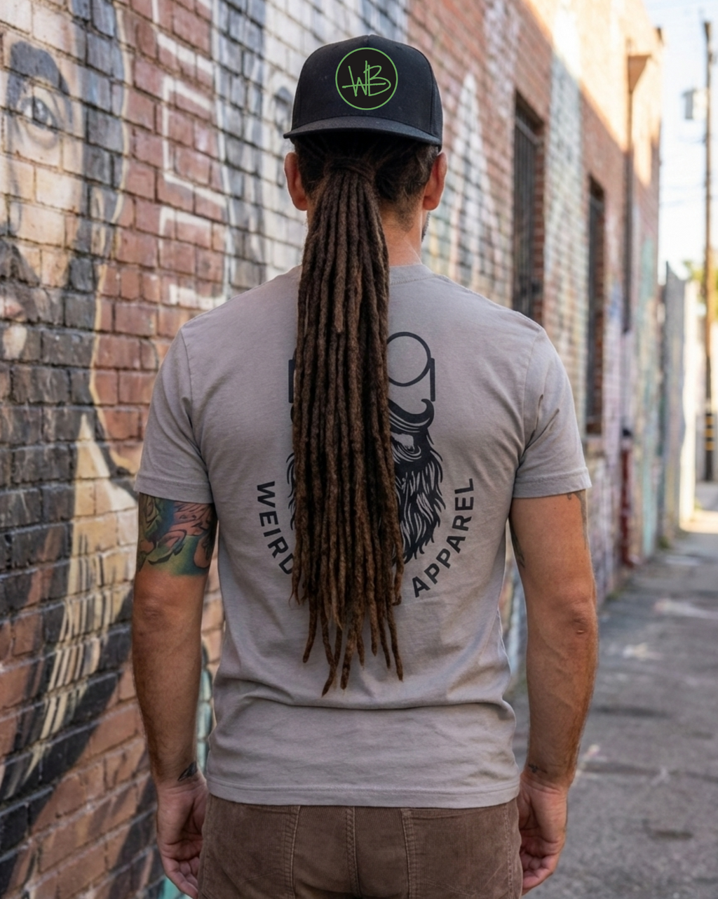 Person wearing a gray t-shirt with a logo and text, standing in an alleyway with graffiti. beard and monocle